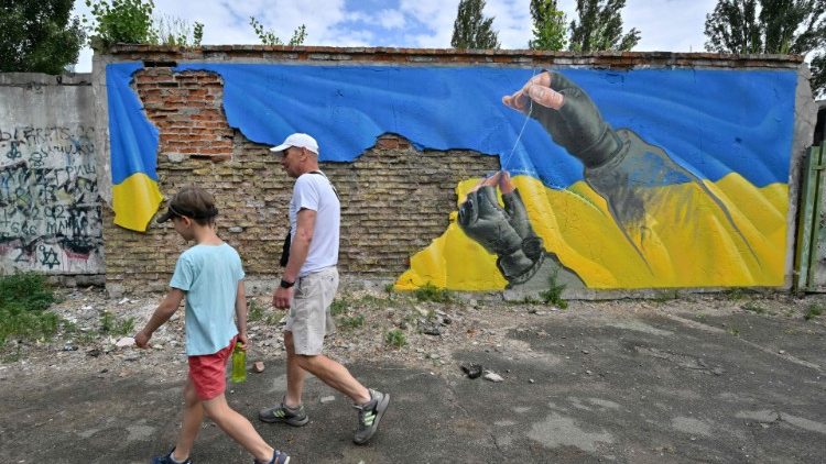 Um homem e seu filho passam por um mural do artista de rua Sasha Korban representando as mãos de um militar costurando partes da bandeira ucraniana, em Kyiv, em 14 de junho de 2022. (Foto de Sergei SUPINSKY / AFP)