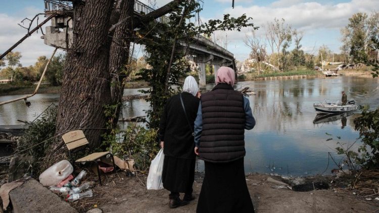 Duas mulheres caminham até um ponto de espera por um barco para cruzar o rio Siverskyi Donets ao lado de uma ponte destruída na cidade recentemente retomada de Sviatohirsk, na região de Donetsk, em 9 de outubro de 2022, em meio à invasão russa da Ucrânia. (Foto de Yasuyoshi CHIBA/AFP)