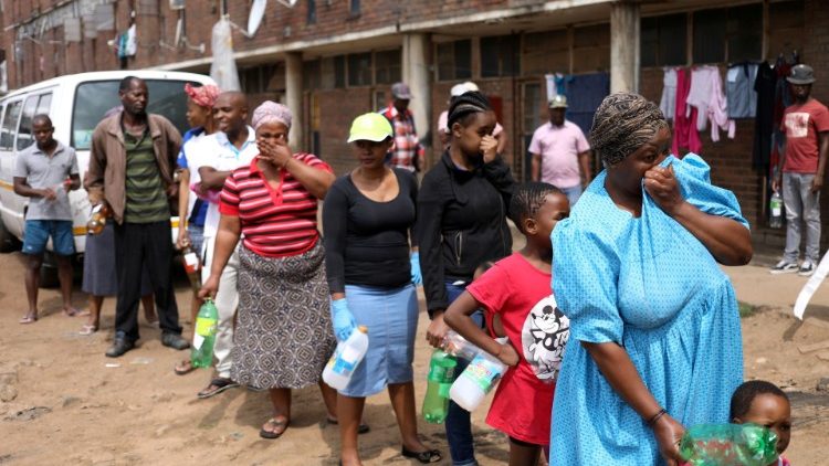 Residentes del Madala Men's Hostel se tapan la nariz mientras esperan en fila para recoger el líquido desinfectante que entregan los trabajadores sanitarios, en Alexandra, Sudáfrica.