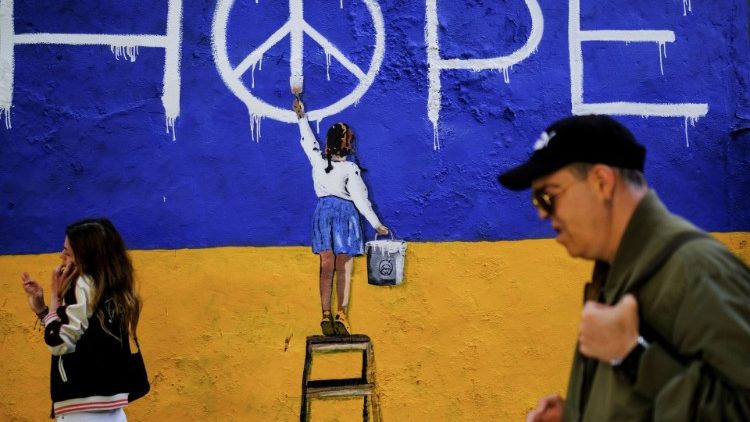 People walk past a mural with the colours of the Ukrainian flag and a girl painting the now "peace" sign