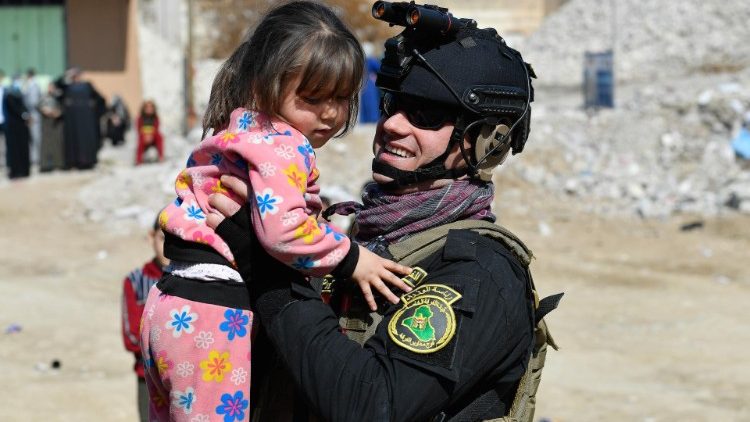 Mosul.  Entre os escombros uma menina olha para um militar com equipamentos de combate que a acaricia, a pega no colo e ela sorri 