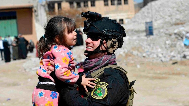 In Mosul, against a backdrop of rubble, a little girl looks at a soldier and smiles as he lifts her up